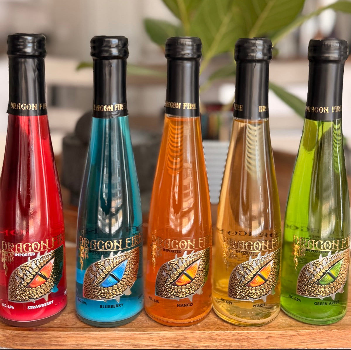 Five Dragon Fire flavored bottles in red, blue, orange, yellow, and green, lined up on a wooden surface with a blurred kitchen background and a green plant.