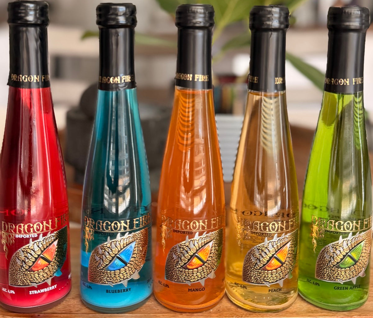 Five Dragon Fire flavored syrup bottles in red, blue, orange, yellow, and green, lined up on a wooden surface with a blurred kitchen background and a green plant.