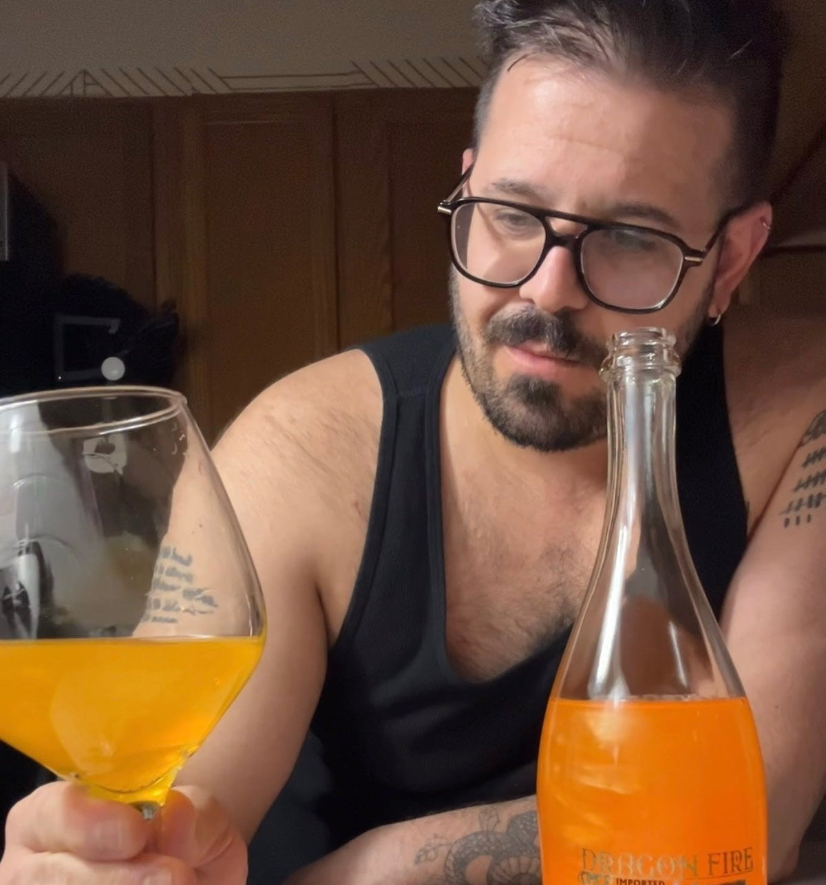 A man wearing glasses and a black tank top holds a stemmed glass filled with an mango dragon fire wine.
