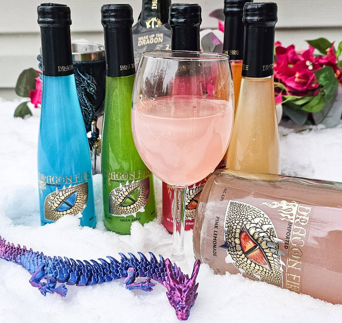 A group of colorful wine and sparkling wine bottles arranged in the snow beside a filled wine glass, with a decorative blue and purple dragon figurine in the foreground. The bottles have black caps and labels featuring a gold and red eye design.