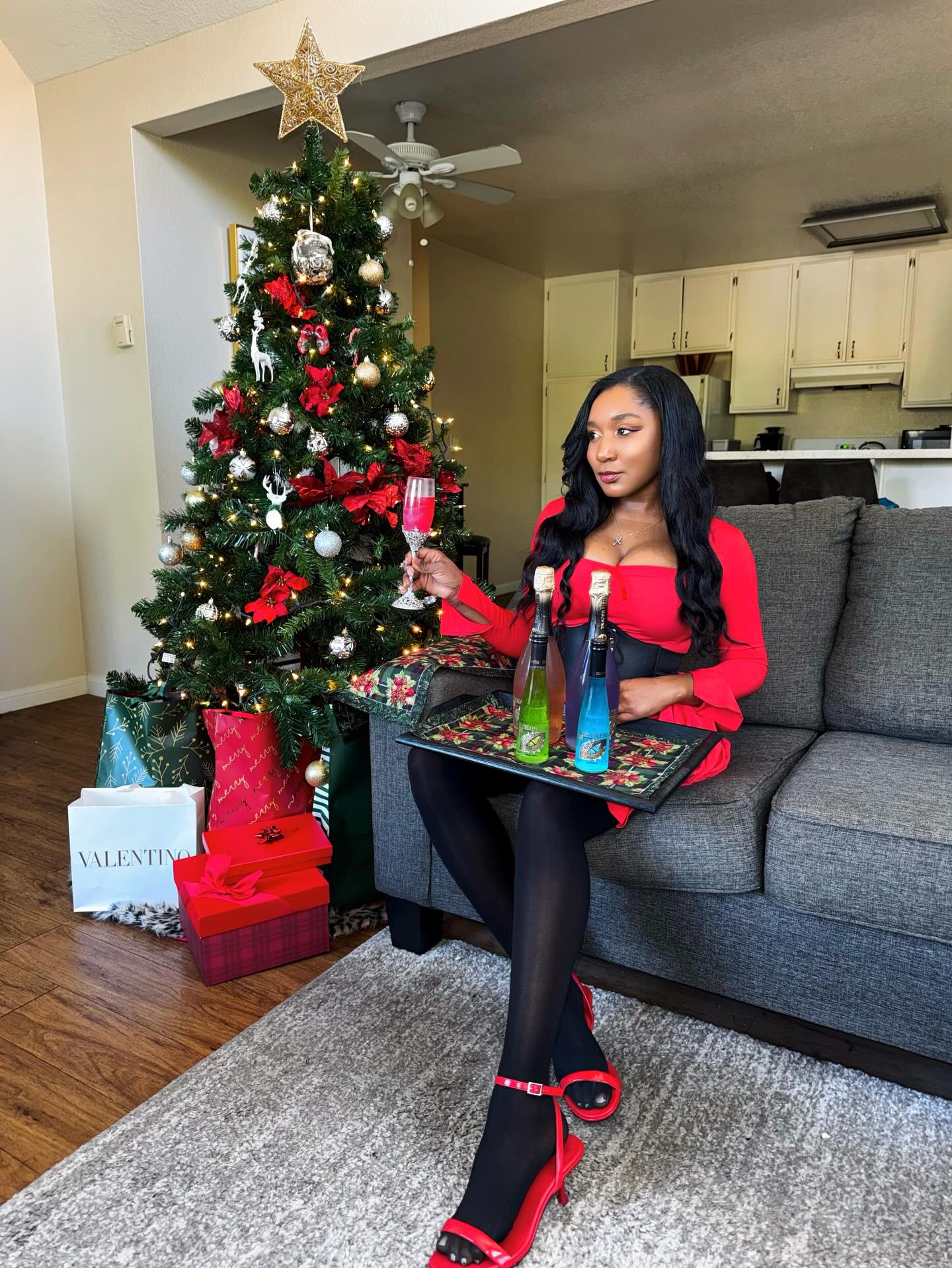 A woman in a red dress and black tights sits on a gray sofa in a living room, holding a tray with bottles and a wine glass. A decorated Christmas tree, wrapped gifts, and a kitchen are visible in the background.