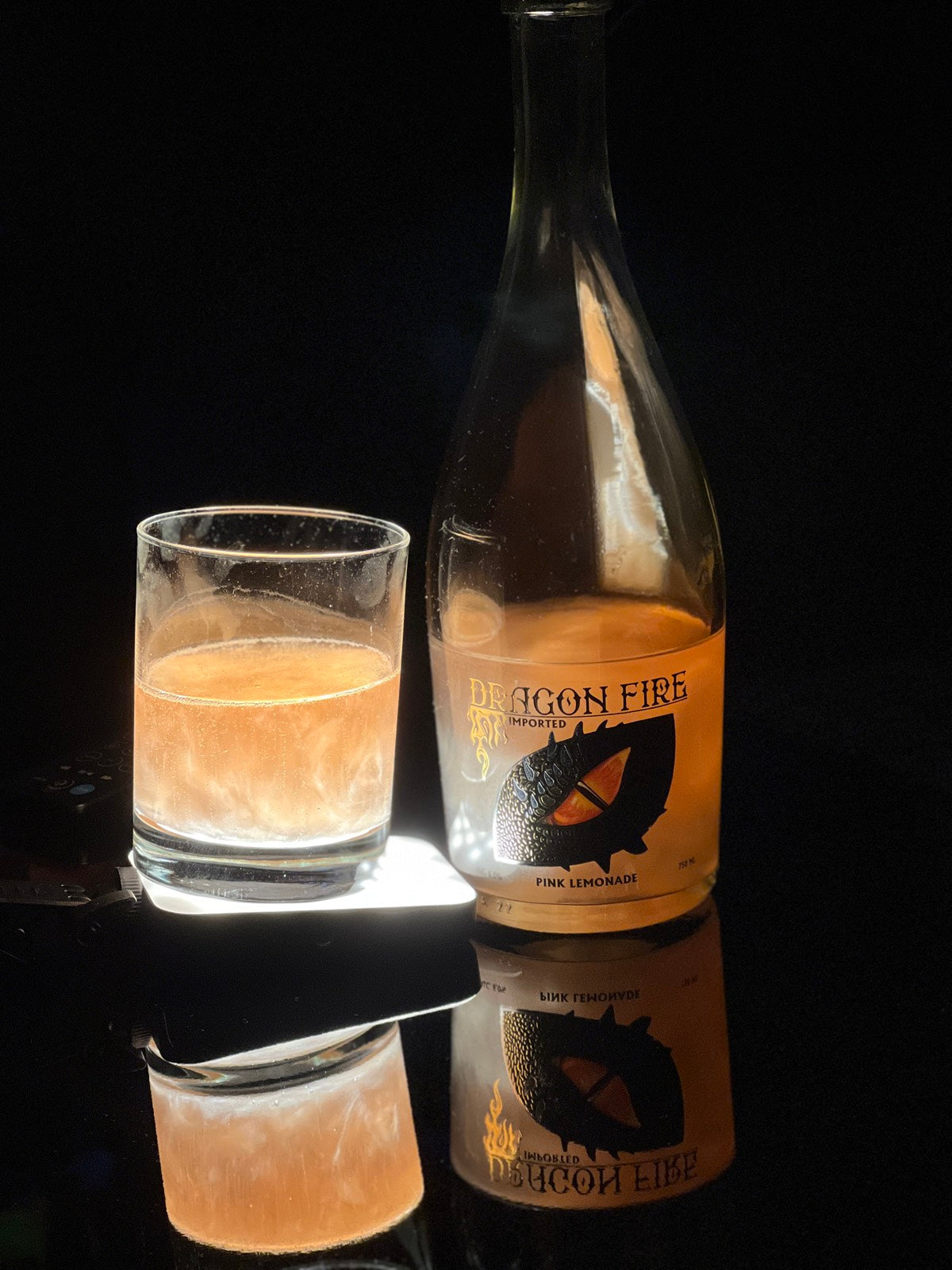 A bottle of Dragon Fire Pina Limescares alongside a clear glass filled with an amber-colored beverage, set against a dark background with a reflective surface showing their reflections.