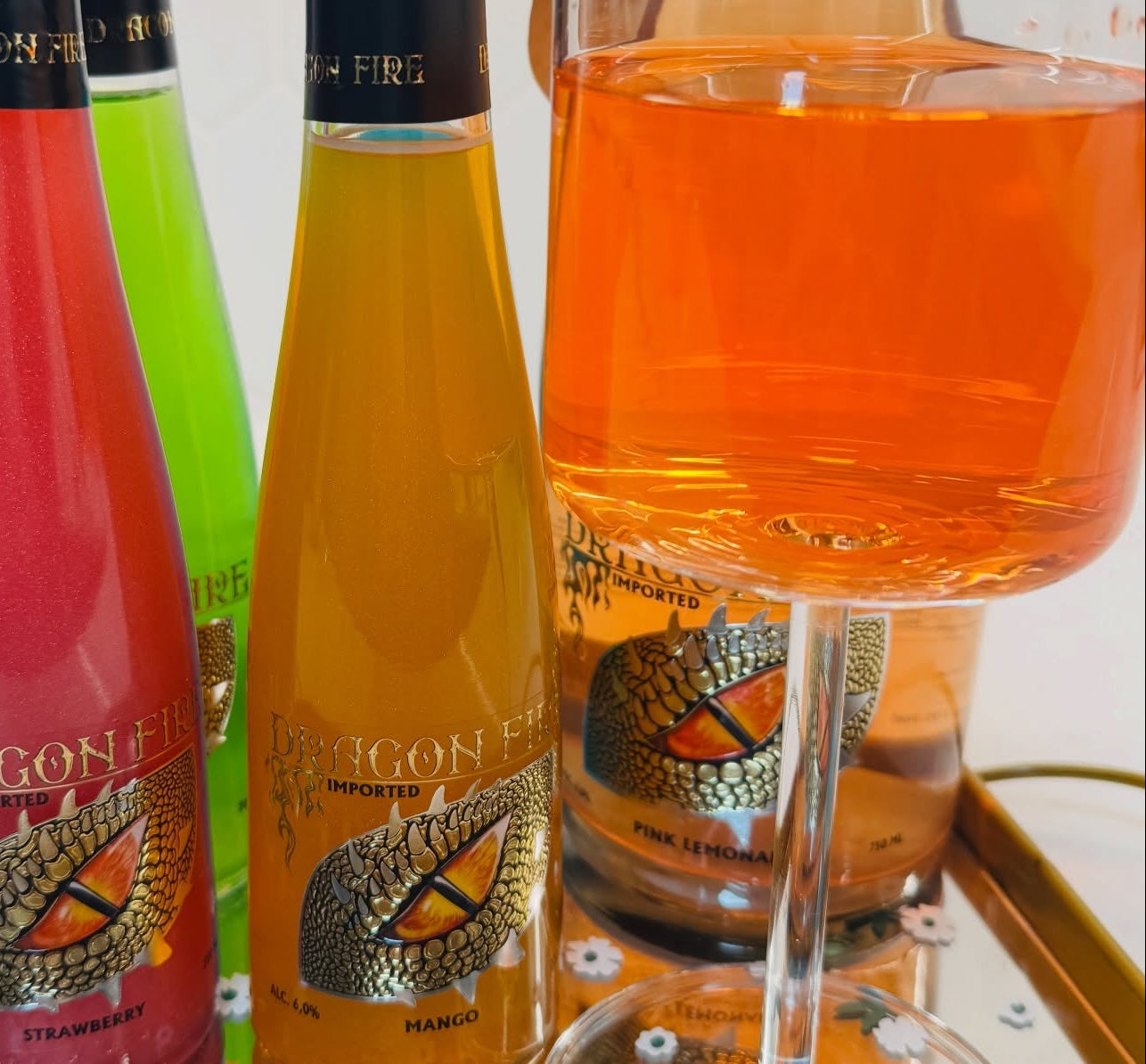 Three Dragon Fire sparkling wine bottles in pink, green, and orange tones, alongside a stemmed glass filled with the orange-colored beverage, set against a light background.